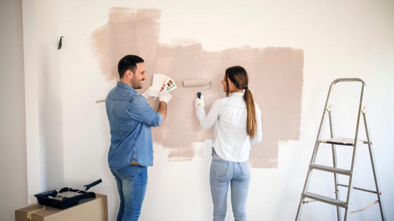 Ein Paar renoviert seine Wohnung, während der Mann eine Farbpalette in der Hand hält und die Frau die Wände mit einer Walze streicht