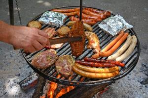 Schwenkgrill mit verschiedenen Fleischwaren, eine Hand wendet Fleisch