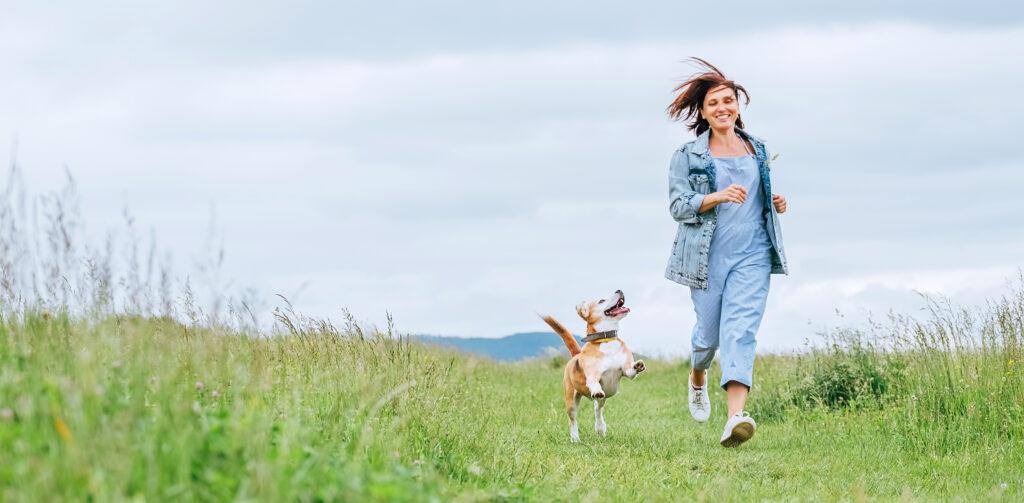 Fröhliche, lächelnde, joggende Frau mit flatternden Haaren und ihrem Beagle-Hund, der mitläuft und ihr auf die Augen schaut.