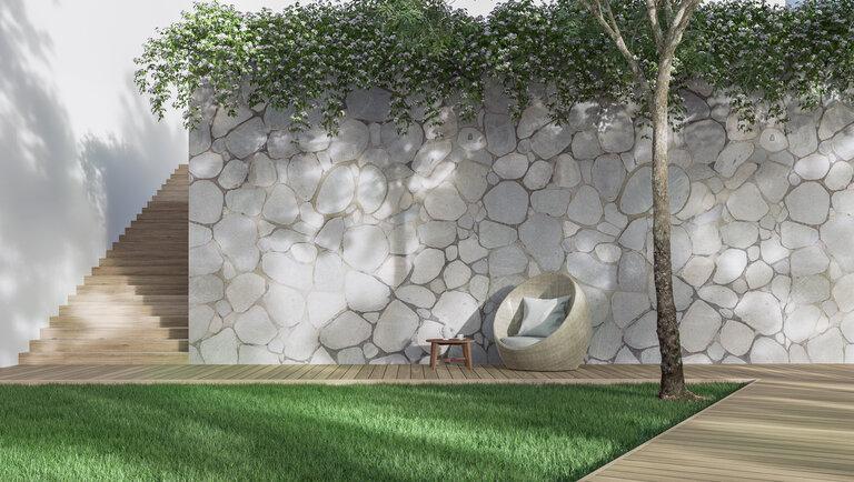 Moderner Garten mit leerer Natursteinmauer in 3D-Putz, mit Holzsteg. Grüner Rasen, große Bäume spenden Schatten.